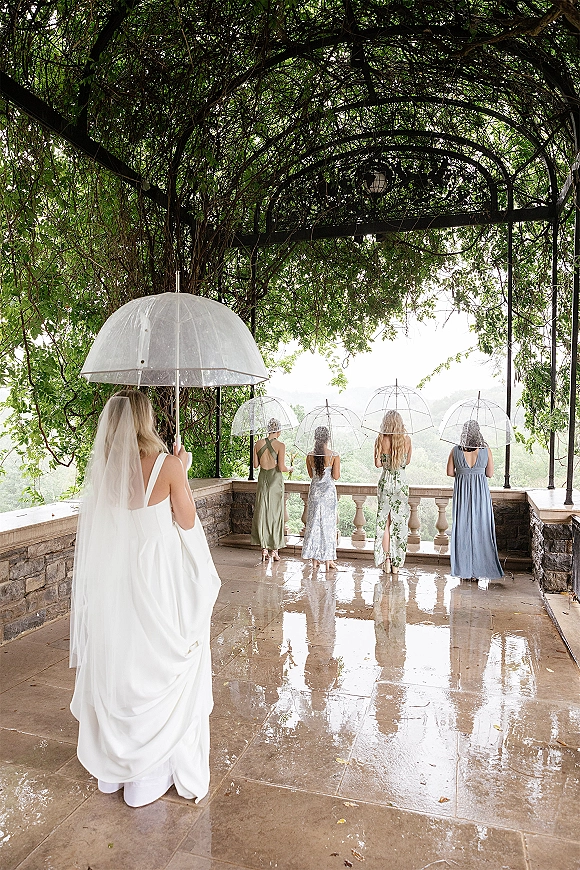 Bridesmaid photo of bridesmaids holding umbrellas as the veiled bride walks toward them on a wet stone terrace under a vine pergola