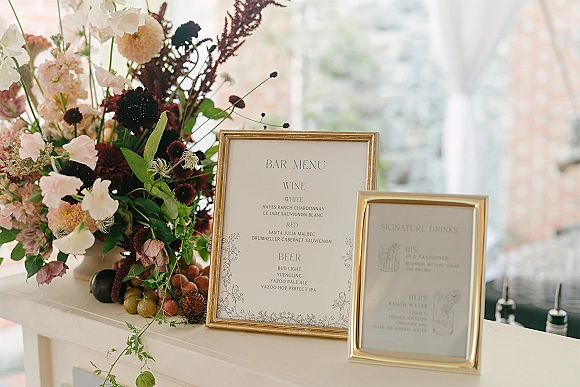 Wedding bar menu in a gold frame beside a framed bar menu sign, with floral arrangement and grapes on a bar counter in window light