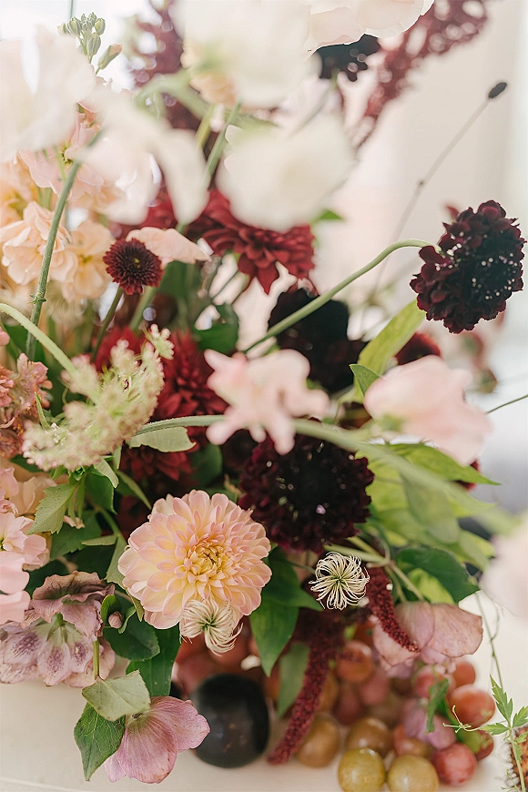 Wedding floral arrangement with fall wedding flowers, blush and burgundy dahlias, greenery, and grapes on a tabletop against white backdrop