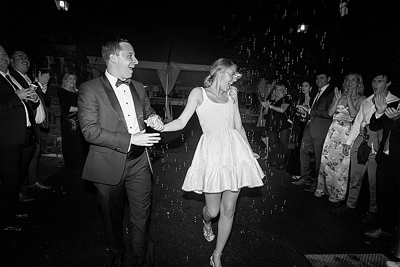 Wedding exit with bride in short reception dress and groom in tuxedo laughing hand in hand through bubbles under string lights at night