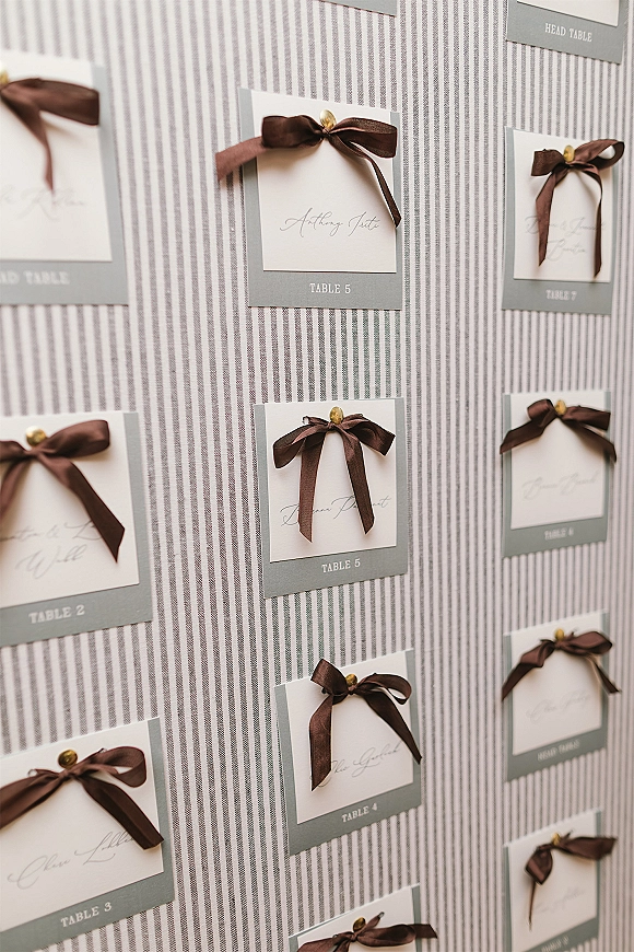Seating chart with gray place cards tied in satin ribbon bows and calligraphy names, pinned with brass tacks on a striped fabric backdrop