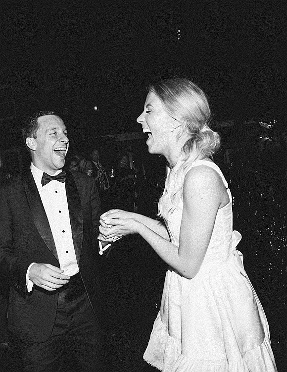 Reception dance moment as bride in a short wedding dress laughs with groom in tuxedo and bow tie on a dark dance floor with guests behind
