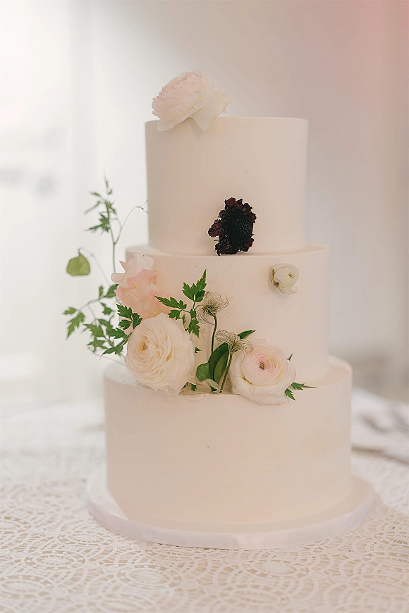 Wedding cake with three tier wedding cake design, smooth white buttercream and fresh flowers on a lace tablecloth by sheer curtains in window light