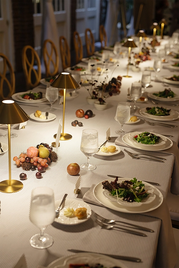 Reception tablescape on a long banquet table with striped tablecloth, brass lamps, greenery centerpieces, place cards, and fruit in a paneled dining room