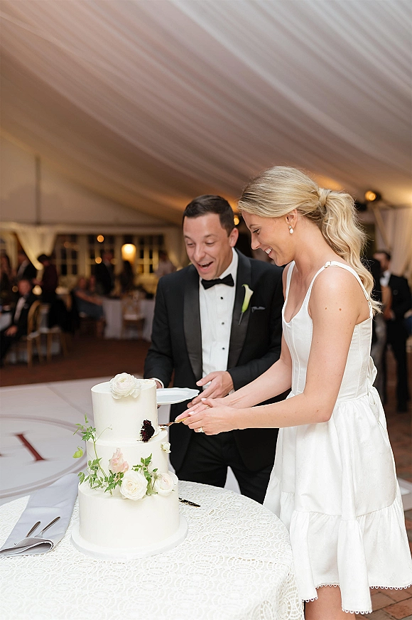 Wedding cake cutting as bride and groom slice a two tier cake with fresh flowers and greenery under bistro lights in a draped tent reception