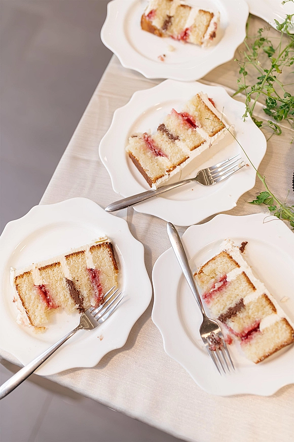 Wedding cake slices on white dessert plates with forks, arranged on a tablecloth with greenery accents in an overhead view