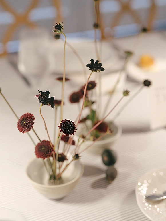 Wedding centerpiece with bud vase centerpiece of dark red and black floral stems on a striped tablecloth, white bowls and plate in soft blur
