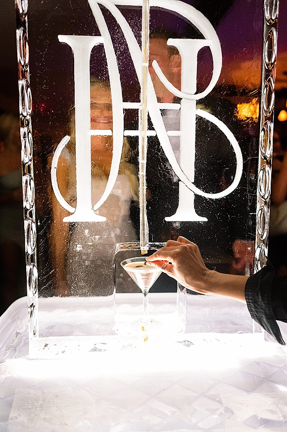 Ice sculpture monogram at a wedding ice sculpture drink luge with martini glass and drip tube on a table, guests in dim reception lighting