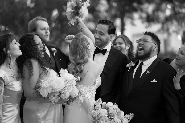 Wedding party photo of the bride and groom with wedding party laughing together, bride holding a bouquet, outdoors with blurred trees behind