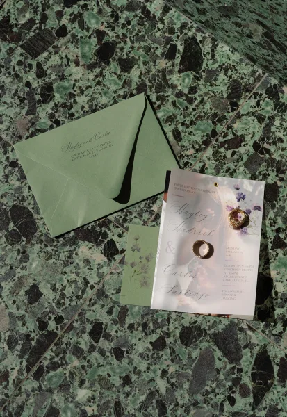 Invitation suite wedding invitation flat lay with sage envelopes, floral cards, wax seal and rings arranged on a green marble surface