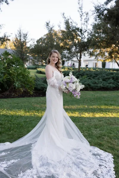 Bridal portrait of a bride holding bouquet of purple and white flowers, looking over her shoulder in a lace gown and long veil on an estate lawn