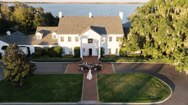 Wedding send off with a bride in gown and veil beside a vintage wedding car on a brick walkway at a white mansion estate drive
