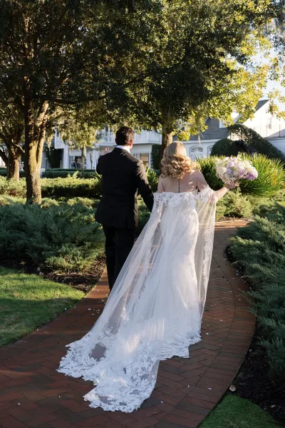 Newlywed walkaway portrait of bride and groom walking away on a brick garden path, her lace train and sheer cape flowing as she holds a bouquet