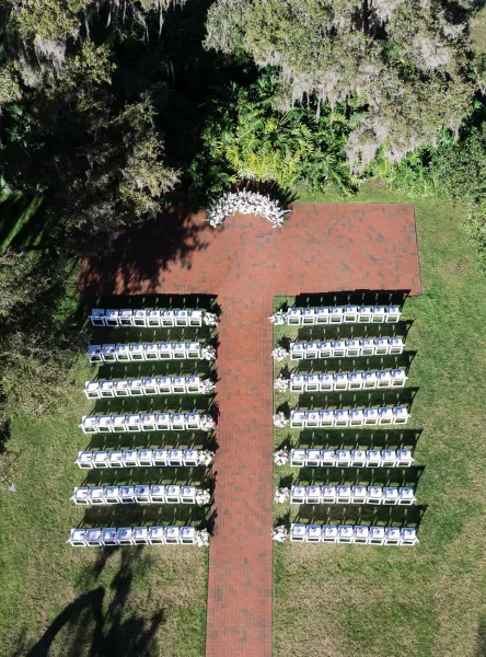 Ceremony setup for an outdoor wedding ceremony with white chairs lining a brick aisle to a floral arch on a lawn beneath mossy trees