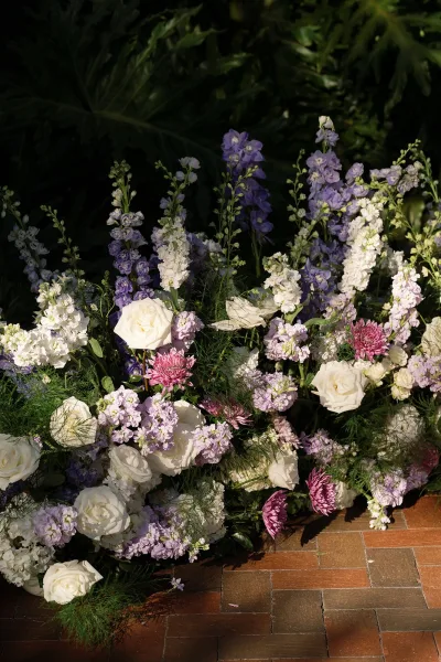 Wedding florals with white roses and purple delphinium in a low ceremony ground flowers meadow along a brick patio in lush garden foliage
