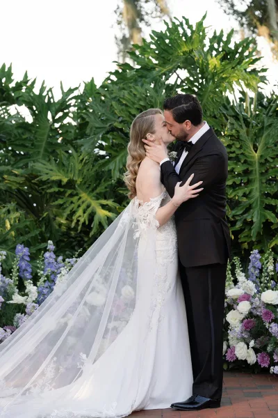 Wedding kiss portrait of bride and groom kissing in a garden, bride in lace dress with long veil as groom in tuxedo holds her face