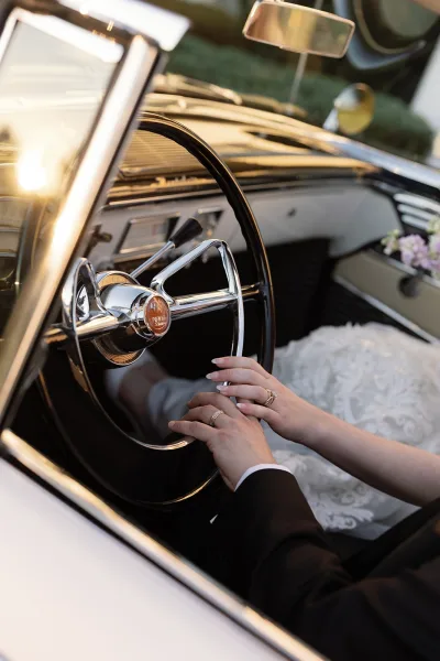 Wedding getaway car interior with bride and groom hands showing wedding rings on the steering wheel, bouquet and lace dress in sunlight