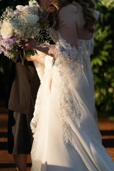 Wedding couple portrait of bride and groom embrace, bride in off-shoulder lace gown holding a white rose bouquet in a golden garden at sunset