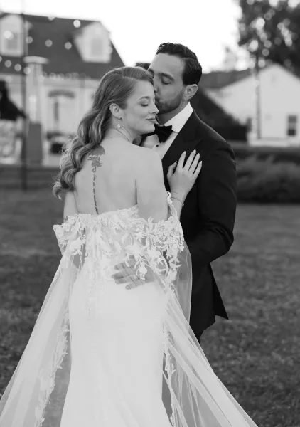 Wedding couple portrait with a forehead kiss wedding photo, bride in off-the-shoulder lace dress and long veil on an estate lawn with string lights