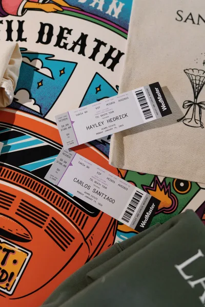 Wedding invitation suite with concert ticket wedding invitations arranged on printed paper and tote bag atop a colorful illustrated poster tabletop flat lay