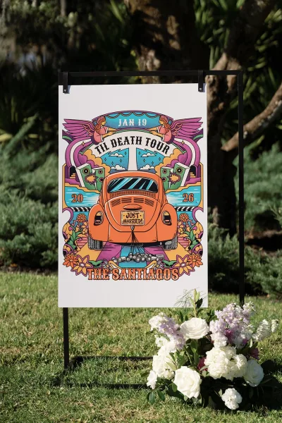 Wedding welcome sign on a black metal stand with white rose floral arrangement, set on a grass lawn with trees and shrubs behind
