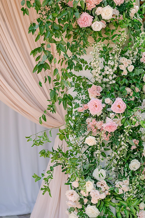 Wedding floral arch with floral arch greenery, pink and white roses, blush blooms, and draped fabric against a white curtain backdrop