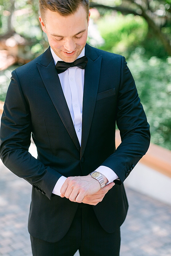 Groom portrait in a black tuxedo and bow tie adjusting his wristwatch on a stone walkway with garden greenery and trees behind him