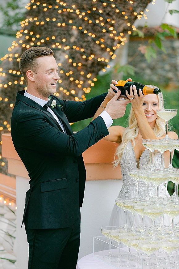 Champagne tower moment as bride and groom pour from a bottle into stacked coupe glasses under string lights on an outdoor patio