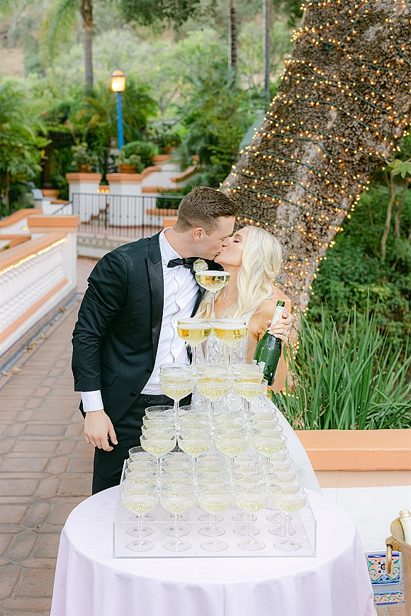 Wedding kiss beside a champagne tower of coupe glasses as the bride holds a bottle, under string lights on an outdoor patio with palms