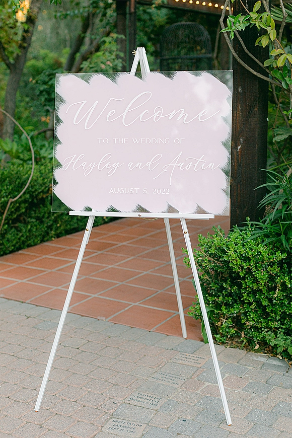 Wedding welcome sign in clear acrylic with white calligraphy on a white easel, set on a brick patio with garden greenery and string lights
