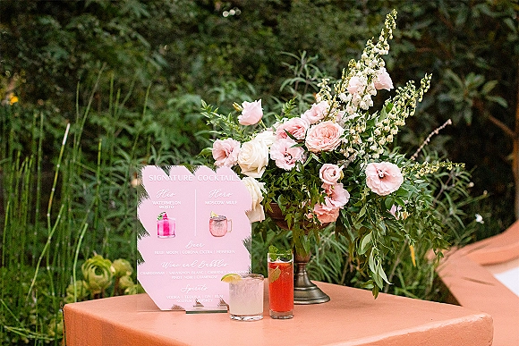 Wedding cocktail sign with signature cocktails sign and pink menu card beside rose and greenery centerpiece, cocktails and glassware on linen table outdoors