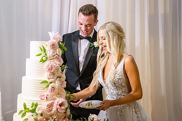 Wedding cake cutting as bride in a beaded gown slices a tiered cake with pink roses and greenery while groom in tux watches by white drape backdrop