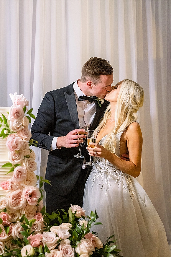 Wedding kiss portrait of the bride and groom kissing with champagne flutes beside a white tiered cake and blush roses against white drapery