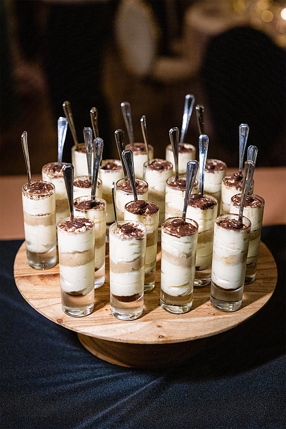 Wedding dessert cups with layered cream and cocoa dusting in glass shooters with mini spoons on a wooden stand in dim reception light