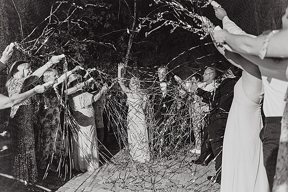 Wedding grand exit with metallic streamers as bride and groom run a cheering tunnel of guests on a brick walkway at night under trees