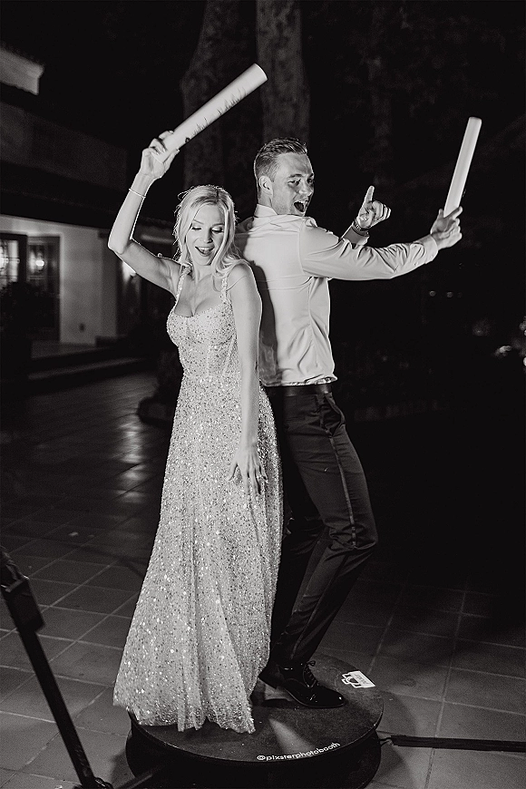 Wedding photo booth couple poses back to back with props and foam glow sticks on a platform at a nighttime outdoor patio reception