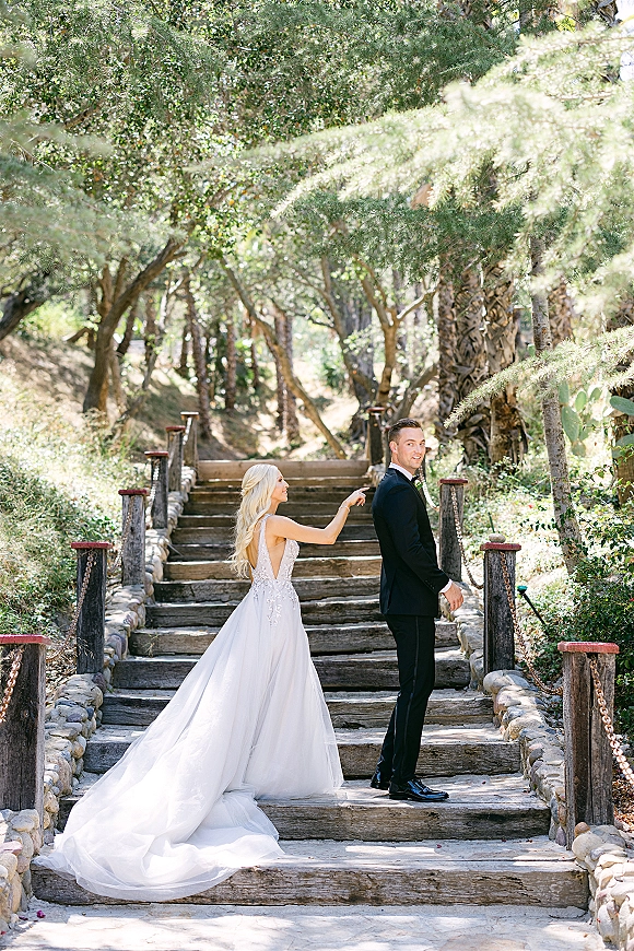 First look moment as bride taps groom’s shoulder on sunlit stone steps, her lace gown train flowing beside his tuxedo boutonniere