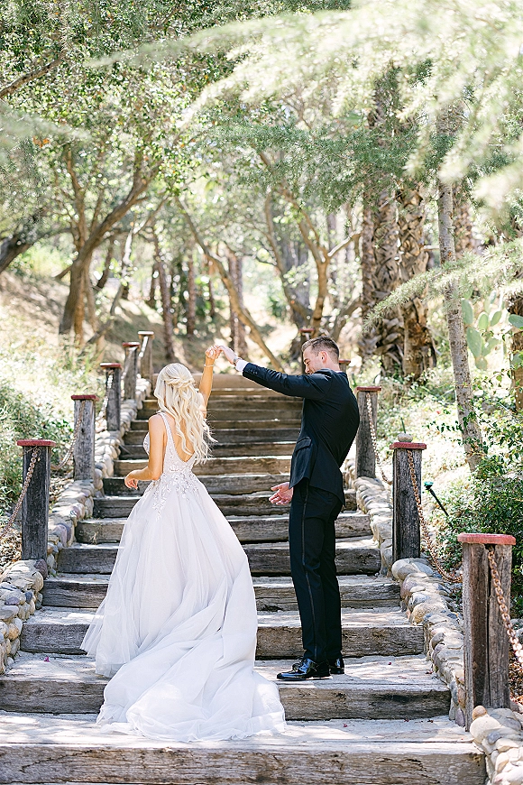 Couple portrait of bride and groom walking away holding hands up wooden stairs, her lace train flowing in sunlit forest path with chain railings