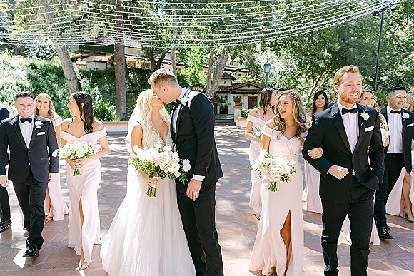 Wedding party portrait of the bride and groom kissing, with bridesmaids and groomsmen holding bouquets in a string-lit courtyard