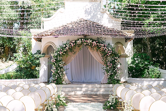 Ceremony setup for an outdoor wedding ceremony with a floral arch and draped fabric, white aisle runner and gold-framed chairs under string lights in a garden courtyard