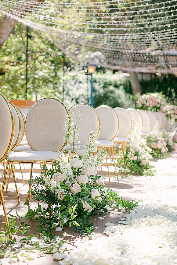 Ceremony aisle decor with white floral ground arrangements, blush roses and greenery garland, gold chairs, rose petals, and string lights outdoors