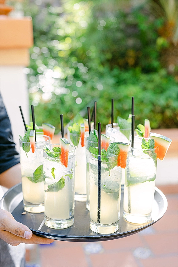 Signature cocktails on a serving tray with glass tumblers, black straws, mint, ice, and watermelon wedges on a green patio backdrop