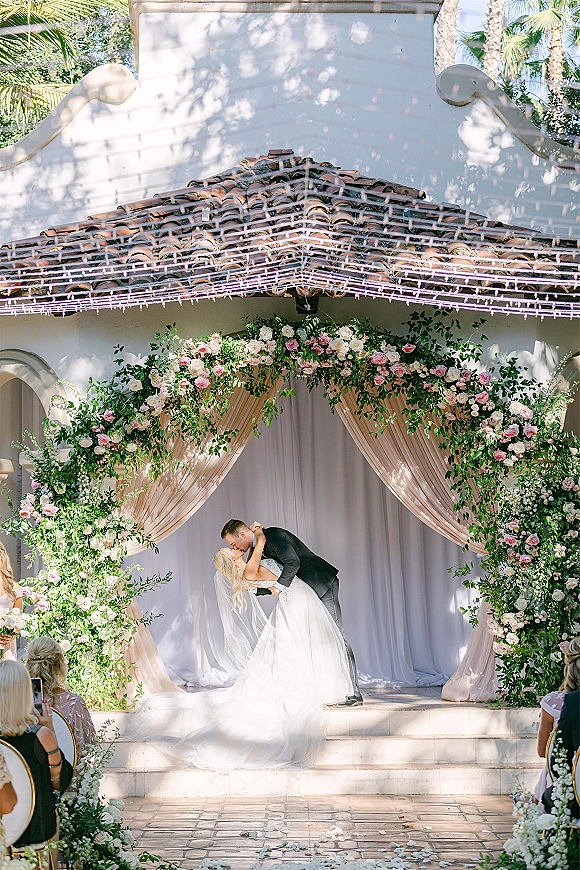 Wedding kiss as the groom dips the bride in a romantic ceremony kiss moment beneath a rose and greenery arch in a sunny courtyard setting