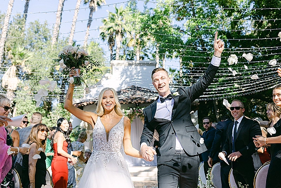 Wedding recessional as bride and groom exit down a garden aisle under string lights, guests tossing rose petals while she raises her bouquet