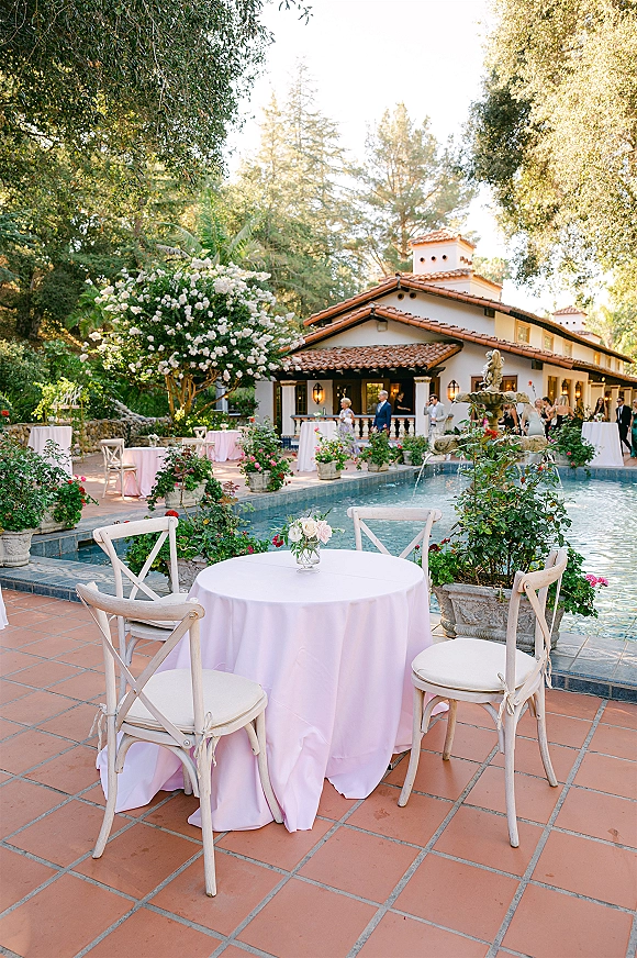 Outdoor reception decor with a round cocktail table in pale pink linens, white rose bud vase centerpiece, and pool fountain on a villa patio