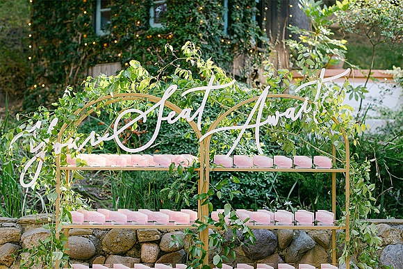 Wedding escort card display with a gold shelf and acrylic calligraphy sign, pink cards and greenery garland against a stone wall and string lights
