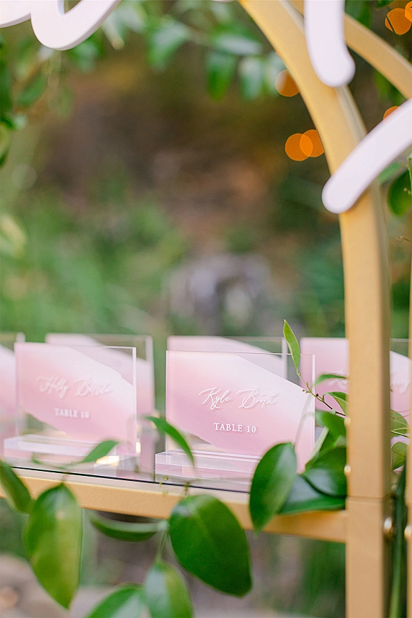 Escort card display with acrylic escort cards in pink calligraphy on a gold frame and glass shelf, greenery vines, garden bokeh lights
