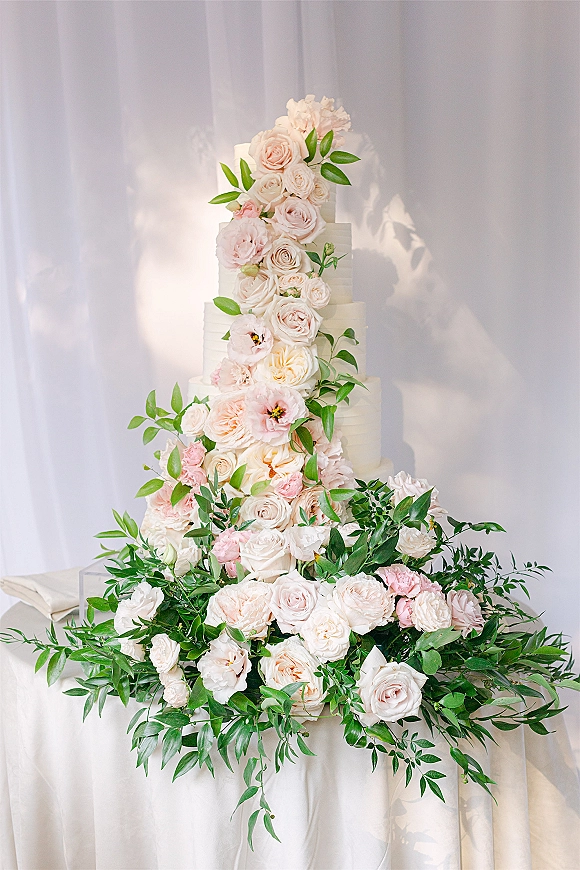 Wedding cake with tiered wedding cake design, blush and ivory roses and greenery cascading on a white table before drapery backdrop