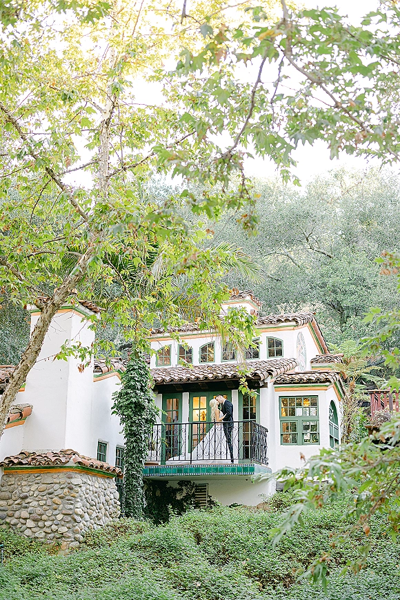 Wedding kiss portrait of bride and groom balcony moment, her long veil draped over iron railing at a stucco villa with greenery hillside backdrop