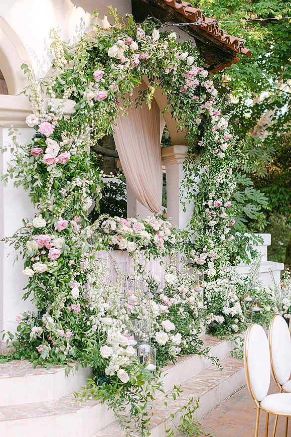 Wedding ceremony arch with a floral wedding arch of pink and white roses, draped fabric, and candles on stone steps at a stucco archway patio